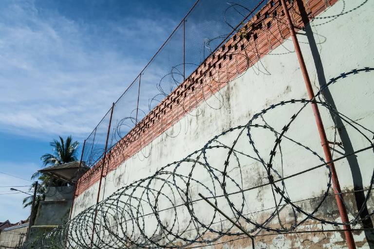 Barbed wire atop a high brick and concrete wall under a blue sky.